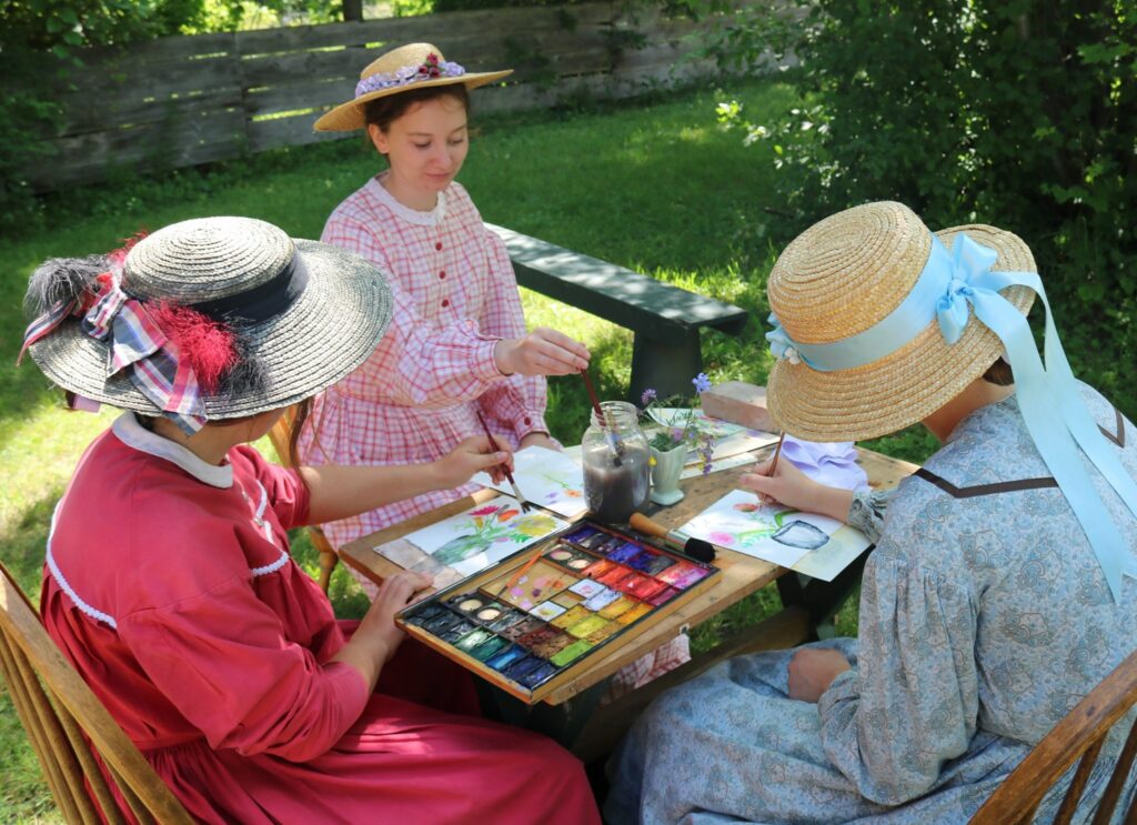Three female dressed in 1860s dresses wearing hats sitting and painting